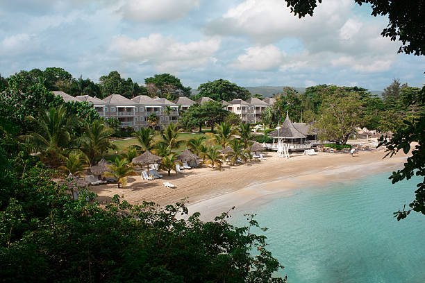 A horizontal photograph of a tropical beach resort in Jamaica, with hotel blocks overlooking a white sand beach, palapa huts and palm trees.